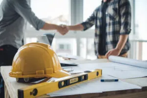 Yellow hard hat and blueprints on a desk in a construction office, with two people shaking hands in the background, symbolizing a M&A targeted acquisition deal.